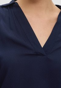 Marineblaues Blusenshirt mit V-Ausschnitt, weichem Griff und subtiler Glanzoptik. Der Stoff fällt sanft um den Ausschnitt und weist gestickte Details auf.