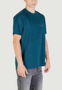 T-shirt in cotone color teal con colletto rotondo, maniche corte e una stampa discreta sul petto; abbinato a jeans grigi chiari.
