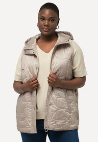 Beige Wattierte Weste mit Kapuze, aus gestepptem Stoff, mit Reißverschluss und Druckknöpfen. Getragen über einem cremfarbenen kurzärmeligen Oberteil.