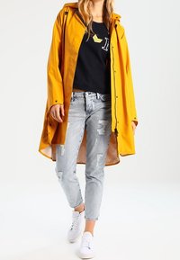 Gele waterdichte regenjas met capuchon, gedragen over een zwart shirt en versleten lichtblauwe spijkerbroek, gecombineerd met witte sneakers.