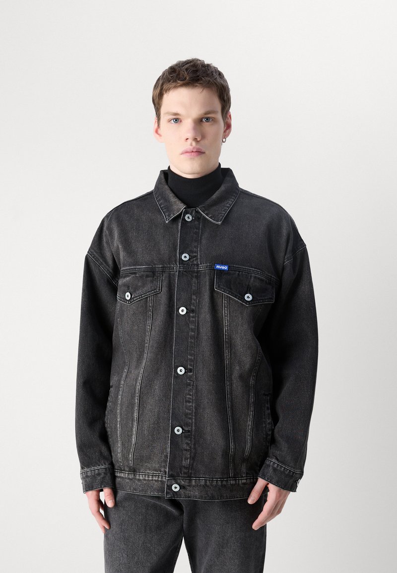 Czarna kurtka dżinsowa o jednolitym kolorze, z zapięciem na guziki, dwiema kieszeniami z przodu i małym niebieskim logo na klatce piersiowej.