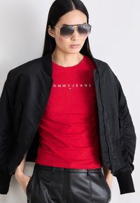 Černá bomber bunda s žebrovanými manžetami, červená košile s nápisem "TOMMY JEANS" a černé kožené kalhoty. Model má na sobě šedé sluneční brýle.