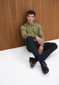 Vihreä pitkähihaine paita, kapea kaulus, kolme nappia. Laivastonsiniset räätälöidyt housut, mustat kengät vaakasaumalla. Puiset paneelit taustalla.