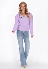 Lavendel gestrickte Strickjacke mit V-Ausschnitt, Knopfverschluss und floralem Stickerei auf den Schultern, kombiniert mit hellblauen Schlagjeans.