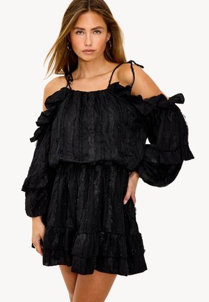 Frau trägt ein schwarzes schulterfreies Kleid mit Puffärmeln und Rüschendetails, stehend vor einem schlichten weißen Hintergrund.