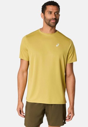 Hombre con camiseta deportiva de manga corta color mostaza y pantalones cortos deportivos verde oliva, de pie y mirando ligeramente hacia la derecha.