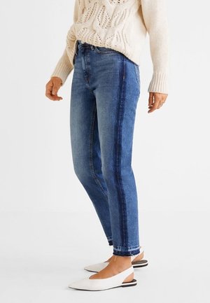 Persona con vaqueros denim azules con una franja lateral oscura, jersey de punto de ochos color crema y zapatos slingback blancos planos, de pie frente a un fondo blanco.