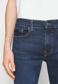 Jeans de mezclilla azul oscuro con bolsillos estándar, costuras naranjas y una etiqueta de cuero negro en la parte trasera. El material tiene una textura suave.