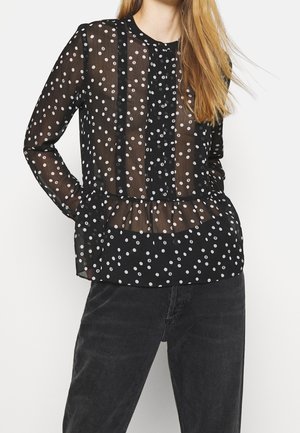 Femme portant un chemisier noir transparent à manches longues avec des pois blancs et un pantalon noir, posant avec les mains dans les poches.