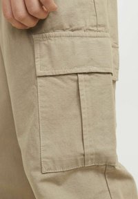 Beige cargo-byxor med en texturerad väv och vertikala ränder samt en stor, platt sidficka säkrad med sömnadskanter.