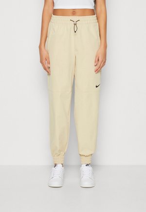 Personne portant un pantalon de jogging beige Nike avec taille et poignets élastiques, associé à des baskets blanches Nike et un top court blanc.