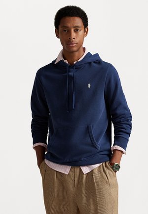 Marineblaue Kapuzen sweatshirt aus weichem Material mit einer Fronttasche, über einer hellrosa blusigen Hemd geschichtet, kombiniert mit karierter Hose.