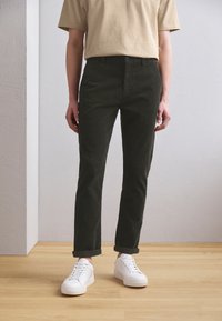 Pantalon en coton vert foncé à coupe droite, doté d'un design classique à cinq poches, associé à des baskets blanches et un t-shirt beige.