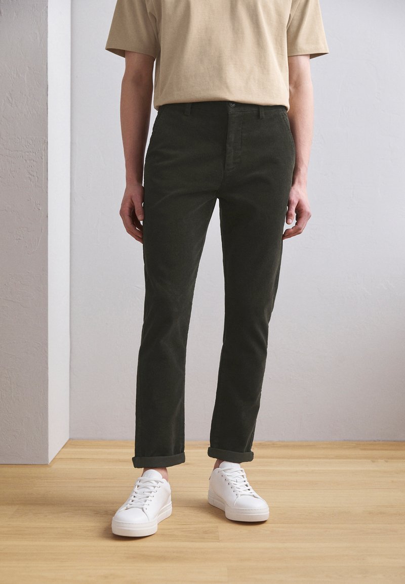 Pantalon en coton vert foncé à coupe droite, doté d'un design classique à cinq poches, associé à des baskets blanches et un t-shirt beige.