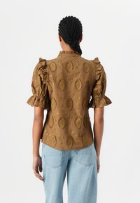 Blusa marrone a fantasia con maniche corte a frange, caratterizzata da un colletto alto e da un intricato ricamo dorato su un tessuto testurizzato.