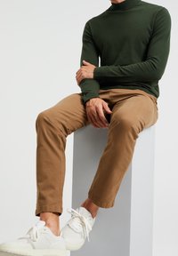 Dark green turtleneck sweater, slim tan pants, and white sneakers. Model seated on a light grey pedestal with a minimalist background.