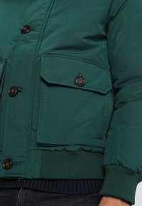 Giacca a vento verde con chiusura a zip e bottone, dotata di due tasche frontali, polsini a coste elastici e tessuto testurizzato.