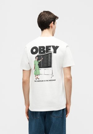 Person trägt weißes T-Shirt mit dem Schriftzug "OBEY" und einer Illustration einer Frau, die wiederholt "I WILL OBEY" an eine Tafel schreibt.