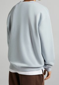 Lichtblauwe sweatshirt met een relaxed fit, ribbelboorden en -zoom, en een zachte stoftextuur. Daaronder is een witte shirt zichtbaar.
