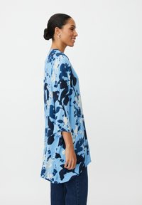 Jasnoniebieska tunika z kwiatowym wzorem w ciemniejszym odcieniu niebieskiego i kremowym. Luźny krój, rękawy do połowy przedramienia, zaokrąglony dół, miękka tekstura, lekka tkanina.