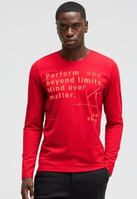 Red long-sleeve t-shirt made of soft fabric, featuring gold text and graphic designs, crew neck, and a slim fit. Text includes motivational phrases.