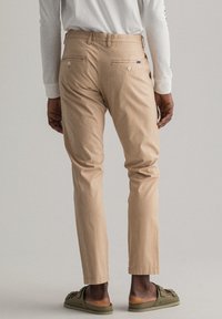 Pantalon en coton beige à coupe slim, présentant une texture lisse, des poches arrière avec fermetures à boutons, et des ourlets retroussés. Porté avec des chaussures vertes sans lacets.