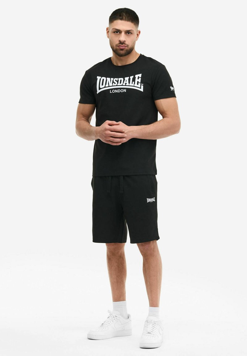 Homme debout, mains jointes, portant un t-shirt noir Lonsdale London, un short noir, des chaussettes blanches et des baskets blanches sur fond blanc.