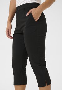 Persoon in zwarte cropped broek met zijsplits aan de zoom en hand in de rechterzak, tegen een effen achtergrond.