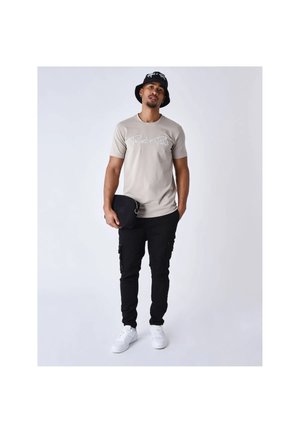 Uomo con t-shirt beige con la scritta "Project x Paris", pantaloni cargo neri, sneakers bianchi, cappello bucket nero, che tiene una piccola borsa nera.