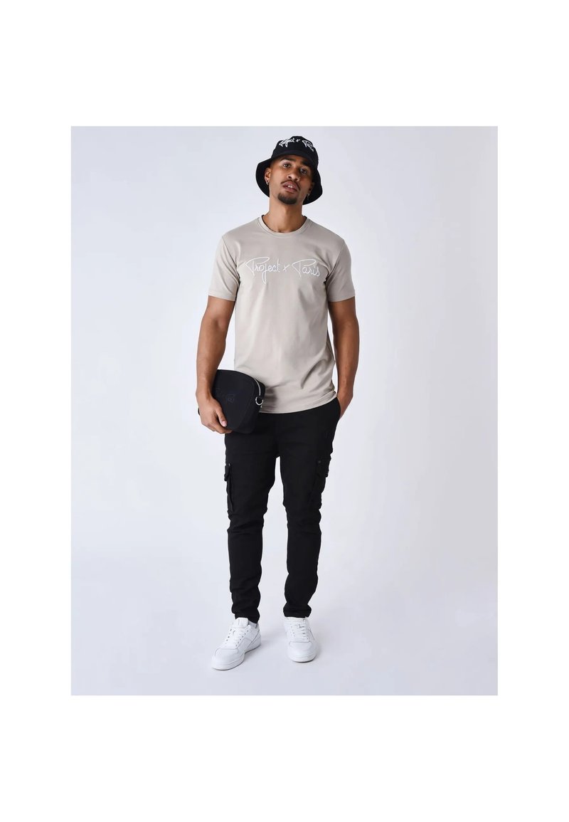 Homme portant un t-shirt beige avec le texte « Project x Paris », un pantalon cargo noir, des baskets blanches, un bob noir, tenant un petit sac noir.