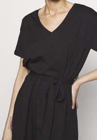 Czarna sukienka do kolan, uszyta z miękkiego materiału, z dekoltem w kształcie litery V, krótkimi rękawami i wiązanym paskiem w talii. Gładka faktura i prosty design.