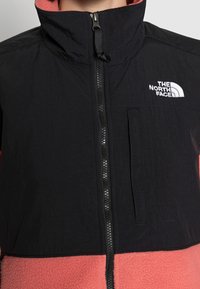 Osoba ubrana w czarną i koralową polarową kurtkę z logo The North Face na klatce piersiowej, z zapięciem na suwak z przodu i wysokim kołnierzem.
