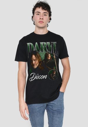 Junger Mann trägt ein schwarzes T-Shirt mit drei Bildern eines Mannes, der eine Armbrust hält, und dem Text „Daryl Dixon“ in Grün und Weiß.
