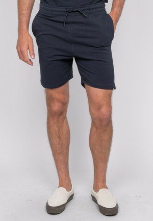Homme portant un short bleu marine à cordon, mains dans les poches, chaussures blanches sans lacets, debout devant un fond uni gris clair.