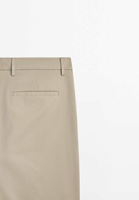 Pantalon beige avec poche passepoilée arrière et passants de ceinture sur fond blanc, montrant une vue partielle arrière de la taille à la partie supérieure de la jambe.