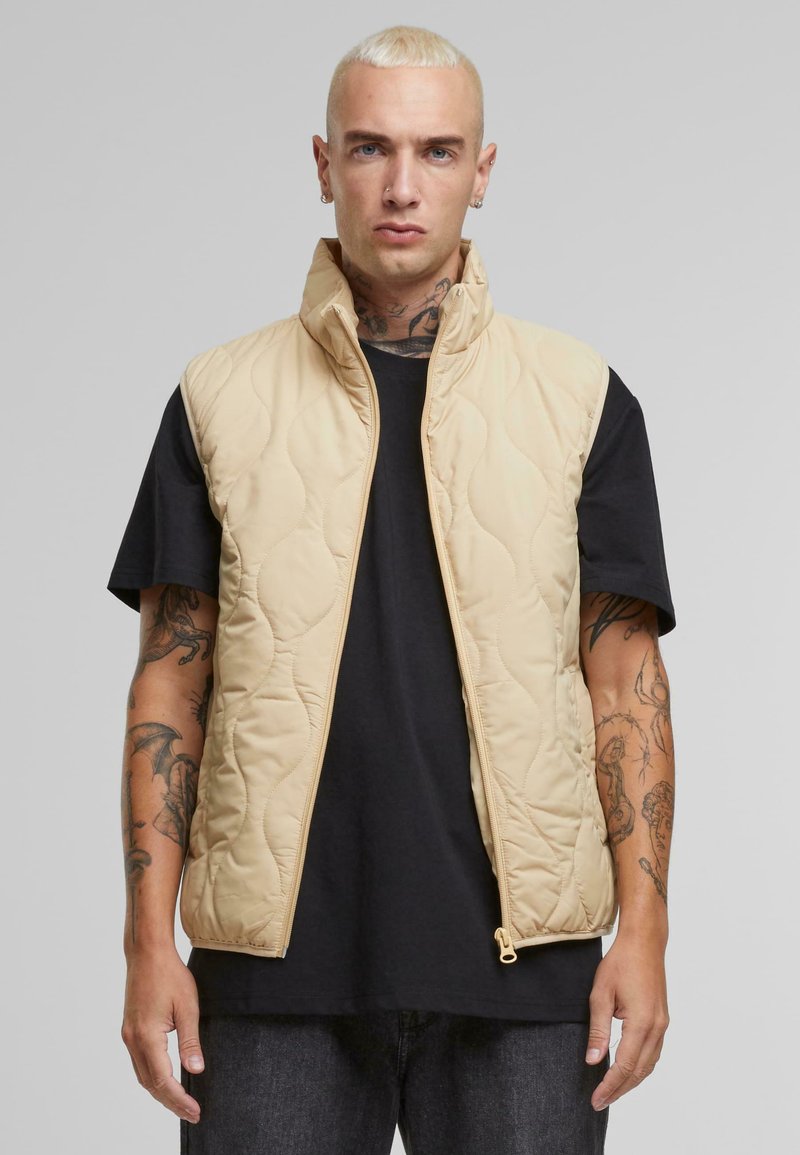 Beige quilted vest with a high collar, featuring a front zipper and lightweight material. Worn over a black t-shirt and dark jeans.