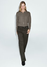 Cardigan marrone con bottoni e texture a costine, abbinato a pantaloni scuri a gamba larga verdi. Stivali neri con punta affusolata completano l'outfit.