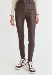Personne portant un pantalon skinny taille haute marron foncé et des baskets montantes blanches avec des accents rouge foncé, debout devant un fond uni.