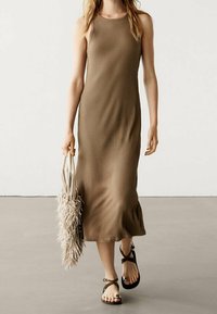 Robe côtelée sans manches, longueur cheville, de couleur taupe, associée à un sac beige à franges et des sandales noires à bretelles croisées.