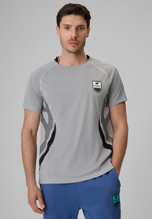 Mężczyzna w szarej sportowej koszulce z czarnymi detalami i niebieskich szortach, stojący z jedną ręką w kieszeni na jednolitym tle.
