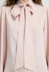 Blusa rosa chiaro realizzata in tessuto liscio, con un colletto alto e un grande fiocco al collo, dotata di maniche lunghe arricciate.