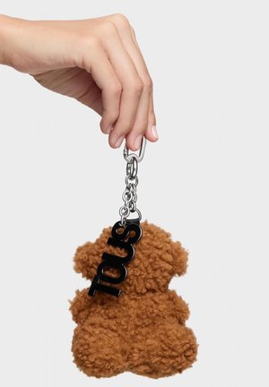 Ręka trzyma brązowy, puszysty brelok w kształcie misia z czarną metką „Tous” na srebrnym łańcuszku na jasnym, jednolitym tle.