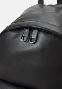 Svart faux skinn ryggsekk med detaljer som inkluderer to glidelåslukkinger med teksturerte trekk, glatt overflate og svak krumming øverst.