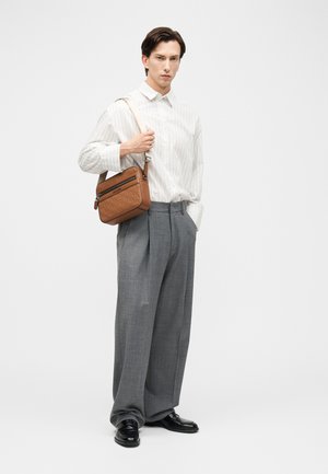 Hombre con camisa blanca a rayas, pantalones grises anchos, zapatos negros, llevando un bolso marrón cruzado sobre el hombro izquierdo contra un fondo blanco.