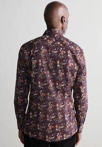 Bloemenpatroon shirt in donker bordeaux met lange mouwen en een klassieke kraag. Kenmerkt zich door verschillende bloempatronen in tinten crème, blauw en bruin.