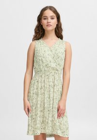 Robe florale verte avec un décolleté en V et sans manches. Taille froncée, jupe fluide et petites fleurs noires sur un fond texturé.