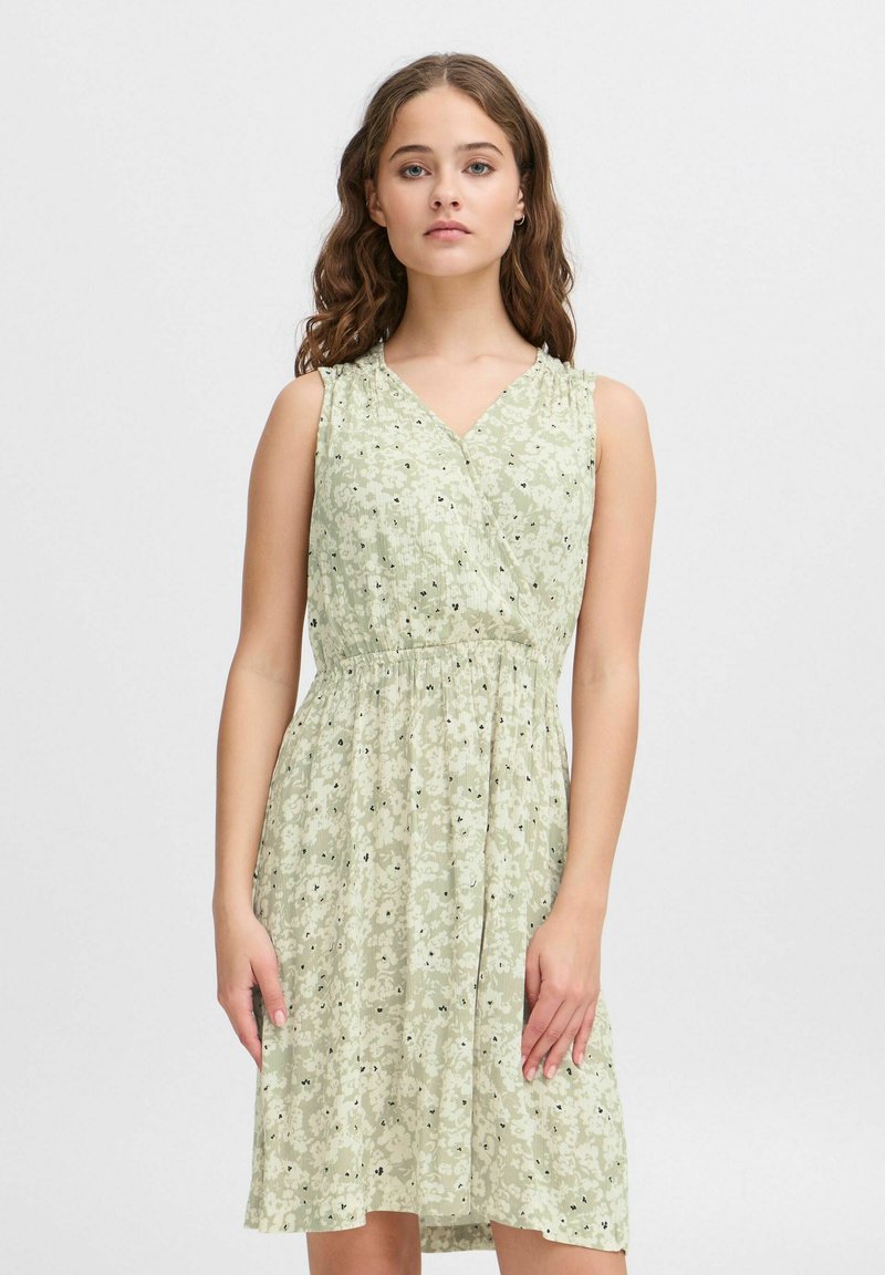 Robe florale verte avec un décolleté en V et sans manches. Taille froncée, jupe fluide et petites fleurs noires sur un fond texturé.