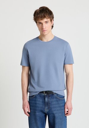 Giovane uomo con capelli castano chiaro che indossa una maglietta azzurro chiaro a maniche corte e jeans blu, in piedi davanti a uno sfondo chiaro e uniforme.