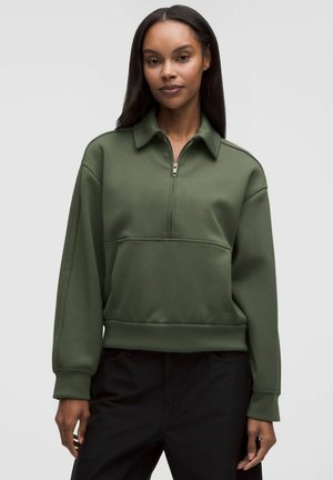 Frau mit langen dunklen Haaren, die einen olivgrünen Pullover mit Reißverschluss und schwarze Hosen trägt, vor einem schlichten hellen Hintergrund steht.