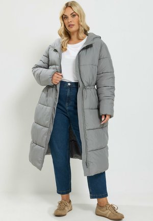 Femme portant un long manteau matelassé gris sur une chemise blanche, un jean bleu et des baskets beiges, debout devant un fond clair uni.